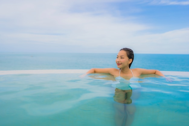 Junge asiatische Frau des Porträts entspannen sich das Lächeln, das um Swimmingpool im Freien im Hotelerholungsort mit Seeozeanblick glücklich ist