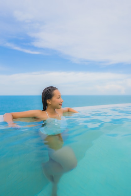 Junge asiatische Frau des Porträts entspannen sich das Lächeln, das um Swimmingpool im Freien im Hotelerholungsort mit Seeozeanblick glücklich ist