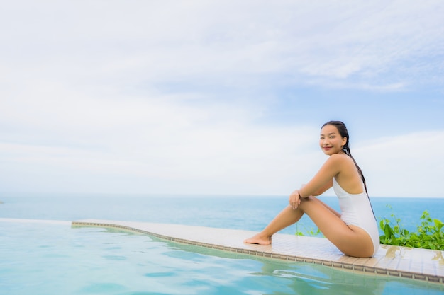 Junge asiatische Frau des Porträts entspannen sich das Lächeln, das um Swimmingpool im Freien im Hotelerholungsort mit Seeozeanblick glücklich ist