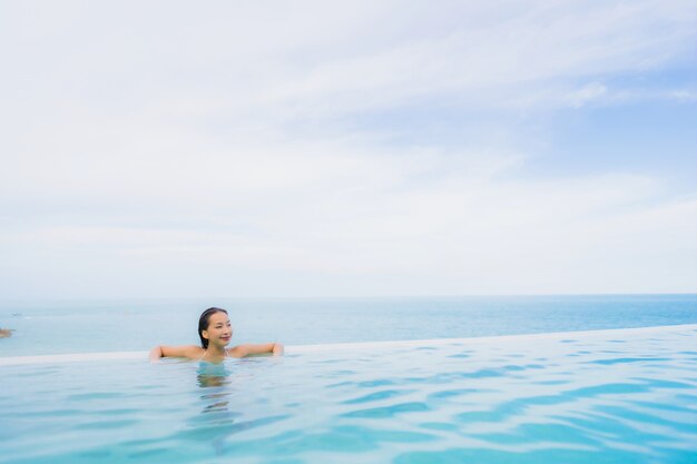 Junge asiatische Frau des Porträts entspannen sich das Lächeln, das um Swimmingpool im Freien im Hotelerholungsort mit Seeozeanblick glücklich ist