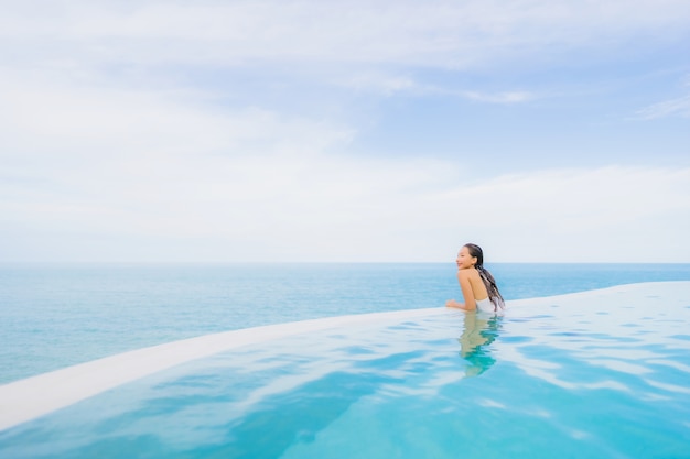 Junge asiatische Frau des Porträts entspannen sich das Lächeln, das um Swimmingpool im Freien im Hotelerholungsort mit Seeozeanblick glücklich ist