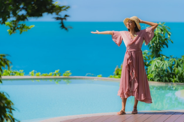 Junge asiatische Frau des Porträts entspannen sich das Lächeln, das um Swimmingpool im Freien im Hotelerholungsort mit Seeozeanblick glücklich ist