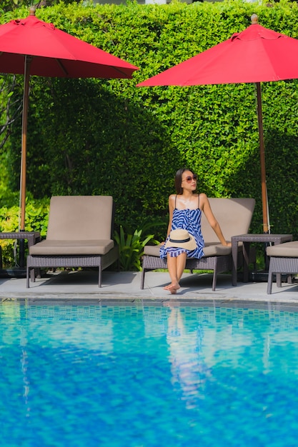 Junge asiatische Frau des Porträts entspannen sich das Lächeln, das um Swimmingpool im Freien im Hotelerholungsort mit Seeozeanblick glücklich ist