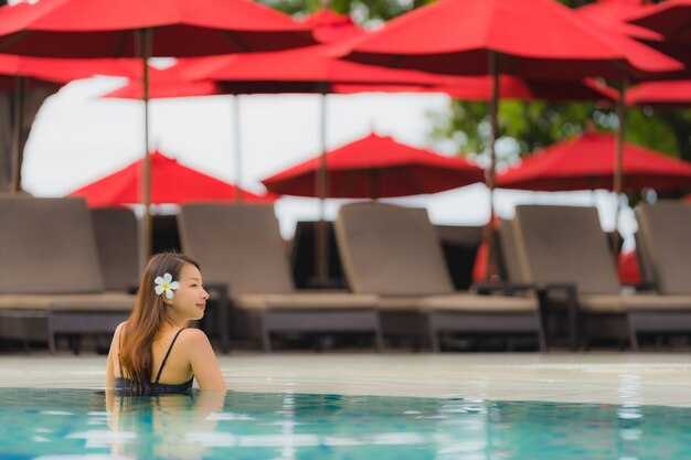Junge asiatische Frau des Porträts entspannen sich das Lächeln, das um Swimmingpool im Freien im Hotelerholungsort mit Seeozeanblick glücklich ist