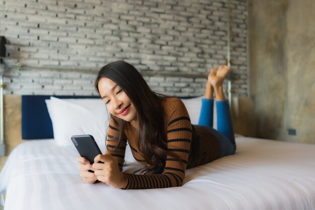 Junge asiatische Frau, die intelligentes Mobiltelefon auf Bett verwendet