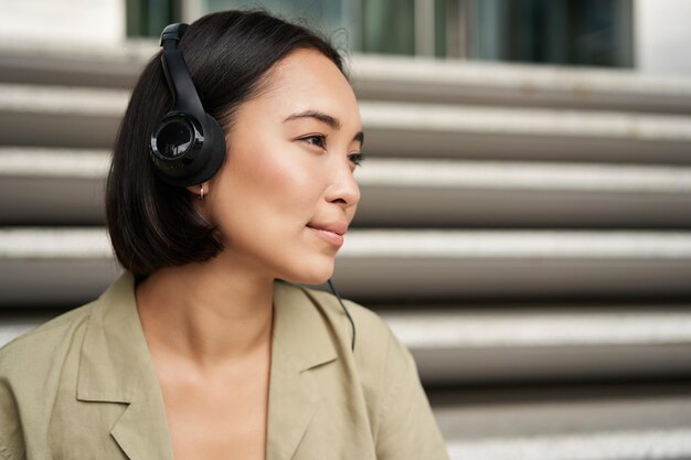 Junge asiatische frau, die musik in kopfhörern hört, die draußen auf der straße sitzen und lächeln