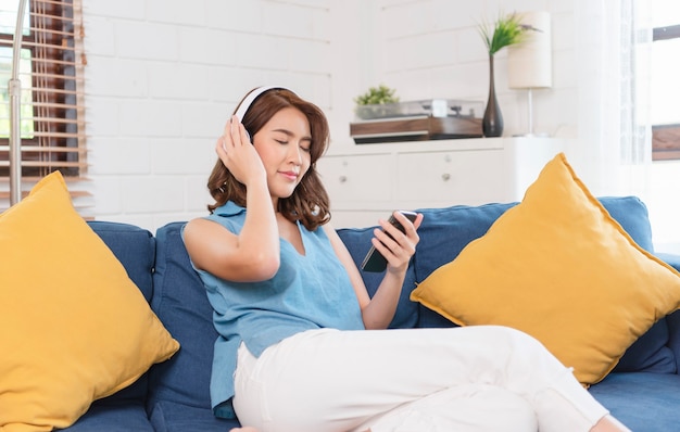 Junge asiatische Frau, die Smartphone verwendet und Musik hört, indem sie eine Verbindung zum Kopfhörer herstellt und am Wochenende auf dem Sofa im Wohnzimmer sitzt.