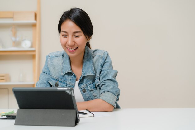 Junge asiatische Frau, die unter Verwendung der Tablette Social Media überprüfend arbeitet, während Sie sich zu Hause auf Schreibtisch im Wohnzimmer entspannen. Zeit zu Hause Konzept genießen.