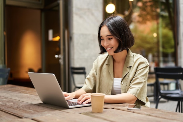 Junge asiatische Frau digitaler Nomade, der fern von einem Café aus arbeitet, Kaffee trinkt und Laptop-Smilin verwendet