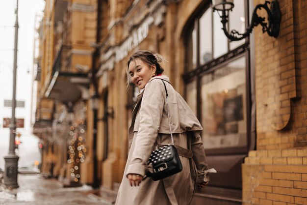 Junge asiatische Frau in Hochstimmung geht durch Stadt im stilvollen Trenchcoat mit kleiner schwarzer Tasche