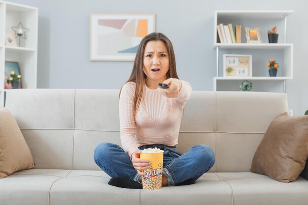 Junge asiatische frau in lässiger kleidung, die zu hause auf einer couch sitzt, mit einem eimer popcorn, der die fernbedienung hält und fernsieht, unzufrieden und verwirrt