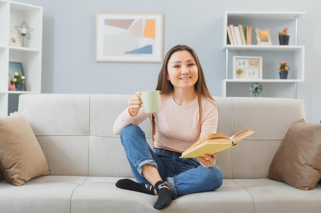 Junge asiatische frau in lässiger kleidung, die zu hause auf einer couch sitzt, mit einer tasse tee, die ein buch liest, glücklich und positiv lächelnd, das wochenende zu hause verbringt