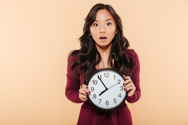 Junge asiatische Frau mit dem gelockten langen Haar, welches die Uhr zeigt fast 8 ist spät oder verfehlt etwas über Pfirsichhintergrund hält