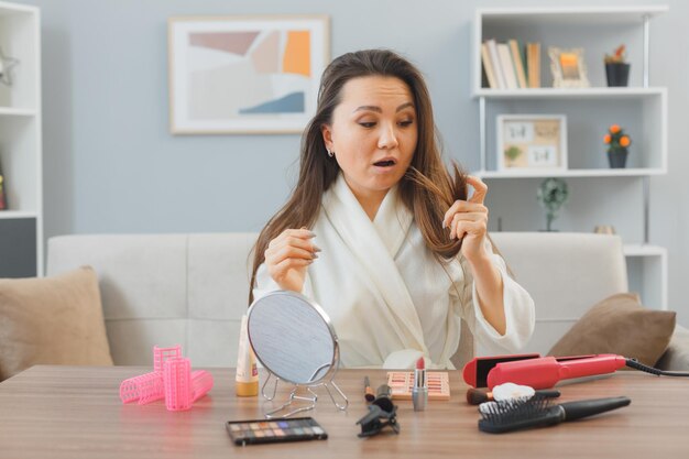 junge asiatische frau mit dunklen langen haaren, die zu hause am schminktisch sitzen und ihr haar betrachten, das sich über die morgendliche make-up-routine aufregt