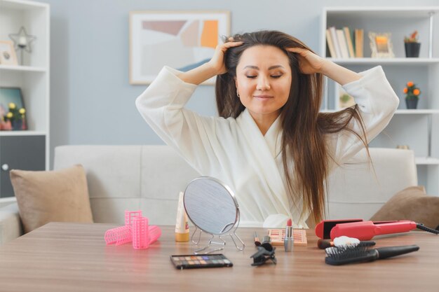 Junge asiatische frau mit dunklen langen haaren, die zu hause am schminktisch sitzt und die kopfhautmassage entspannt, während sie die morgendliche make-up-routine macht