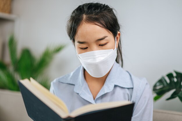 Junge asiatische frau mit gesichtsmaske, die auf dem sofa im wohnzimmer sitzt, sie liest buch während der quarantäne covid-19 selbstisolation zu hause