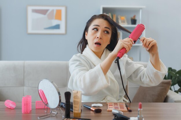 Junge asiatische frau mit handtuch im bademantel sitzt am schminktisch zu hause innen mit lockenstab, der das styling macht, das verwirrt ist, morgens make-up-routine zu machen