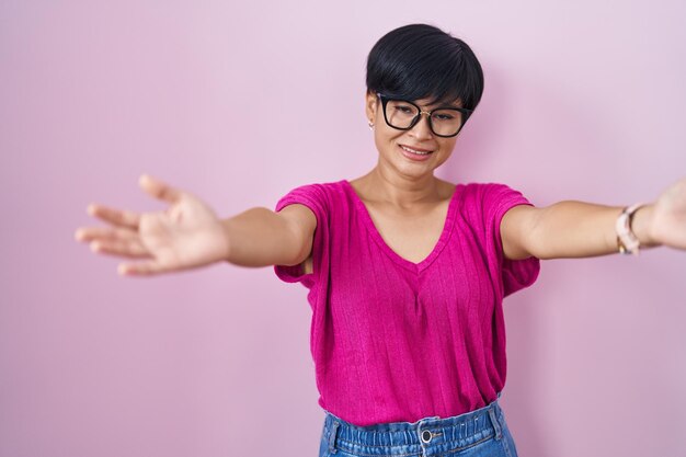 Junge asiatische Frau mit kurzen Haaren, die über rosafarbenem Hintergrund steht und die Kamera ansieht und mit offenen Armen für eine Umarmung lächelt. fröhlicher Ausdruck, der das Glück umarmt.