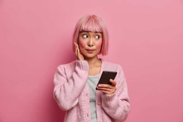 Junge asiatische Frau mit überraschtem Gesichtsausdruck schaut zur Seite, in Freizeitkleidung gekleidet, hält Handy, surft in sozialen Medien, sendet Inhalte, teilt Multimedia online,
