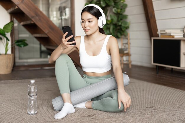 Junge asiatische frau mit wasserflasche hört musik oder podcast in kopfhörern während der fitness zu hause ...