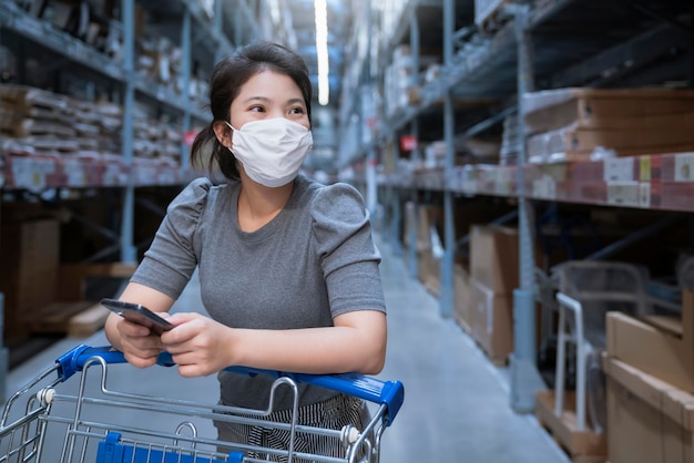 Junge asiatische Frauen in Freizeitkleidung, die mit Einkaufswagen im Kaufhauslager mit neuem normalen Lebensstil einkaufen, tragen Gesichtsmaskenschutz und verwenden Smartphone nach Sperrung von Covid19-Ausbreitung