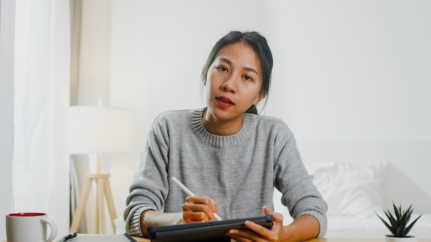 Junge asiatische Geschäftsfrau, die Computer-Laptop verwendet, spricht mit Kollegen über Plan in Videoanrufbesprechung, während sie von zu Hause im Schlafzimmer arbeiten