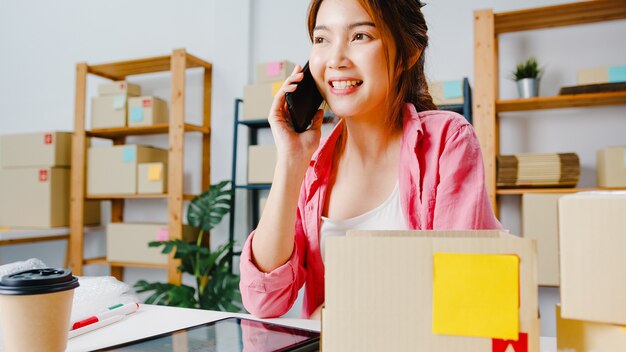 Junge asiatische Geschäftsfrau, die Handyanruf verwendet, der Bestellung empfängt und Produkt auf Lager überprüft, Arbeit zu Hause Büro. Kleinunternehmer, Online-Marktzustellung, freiberufliches Lifestyle-Konzept.