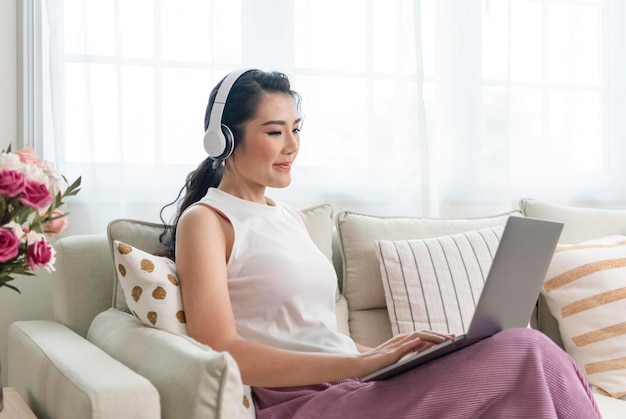 Junge asiatische geschäftsfrau, die laptop-computer verwendet, während sie sich zu hause auf einer couch entspannt, arbeitet von zu hause aus