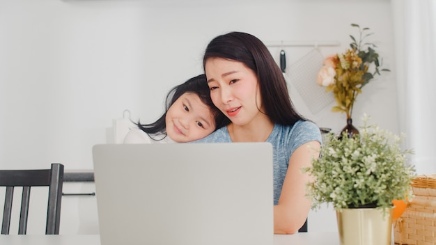 Junge asiatische Geschäftsfrau ernst, Druck, müde und krank beim an Laptop zu Hause arbeiten. Junge Tochter, die ihre Mutter tröstet, die morgens schwer in der modernen Küche am Haus arbeitet.