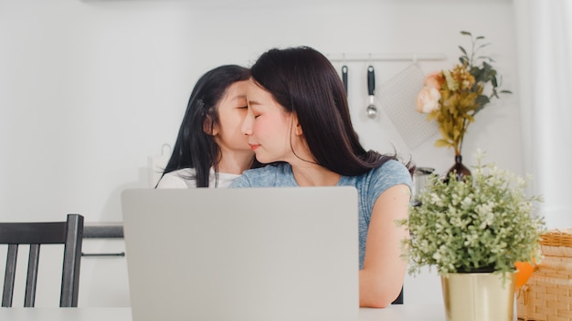 Junge asiatische Geschäftsfrau ernst, Druck, müde und krank beim an Laptop zu Hause arbeiten. Junge Tochter, die ihre Mutter tröstet, die morgens schwer in der modernen Küche am Haus arbeitet.