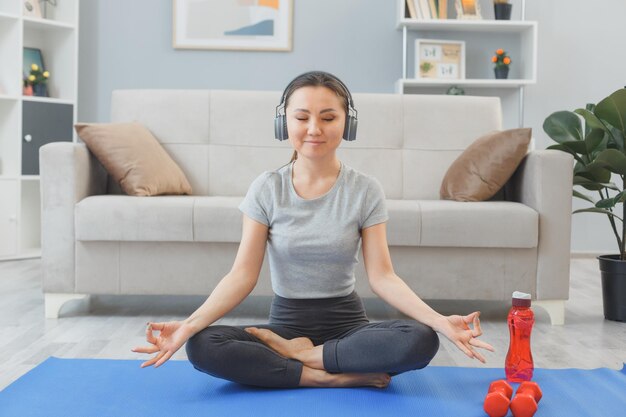 Junge asiatische gesunde Frau mit Kopfhörern, die zuhause zu Hause trainieren und im Wohnzimmer auf einer Yogamatte sitzen und Meditationsgesten machen