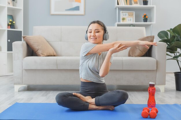 Junge asiatische gesunde frau mit kopfhörern, die zuhause zu hause übungen machen und ihre arme im wohnzimmer ausstrecken, das auf yogamatte sitzt