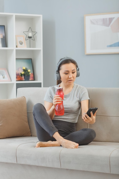 Junge asiatische gesunde frau mit kopfhörern sitzt auf einer couch mit einer flasche wasser zu hause innen müde und überarbeitet nach dem training mit dem smartphone