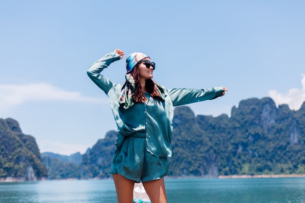 Junge asiatische glückliche Frau Blogger-Tourist in Seidenanzug und Schal und Sonnenbrille im Urlaub reisen um Thailand auf asiatischem Boot, Khao Sok Nationalpark.