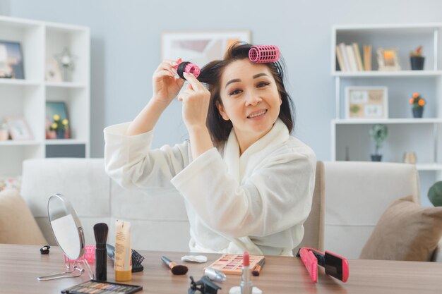 Junge asiatische glückliche frau mit langen dunklen haaren, die am schminktisch zu hause sitzt und haarrollen auf das haar aufträgt, das die morgendliche make-up-routine macht