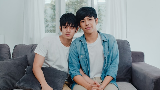 Junge asiatische homosexuelle Paare des Porträts, die glücklich sich fühlen, zu Hause zu lächeln. Asiatische LGBTQ-Männer entspannen sich das toothy Lächeln, das zur Kamera beim morgens liegen auf Sofa im Wohnzimmer schaut.
