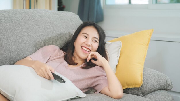 Junge asiatische Jugendlichfrau, die zu Hause, weibliches glaubendes glückliches Lügen auf Sofa im Wohnzimmer fernsieht. Lebensstilfrau entspannen sich am Konzept des Morgens zu Hause.