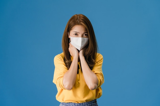 Junge asiatische Mädchen tragen medizinische Gesichtsmaske, müde von Stress und Spannung, schaut zuversichtlich auf Kamera isoliert auf blauem Hintergrund. Selbstisolation, soziale Distanzierung, Quarantäne zur Vorbeugung von Koronaviren.