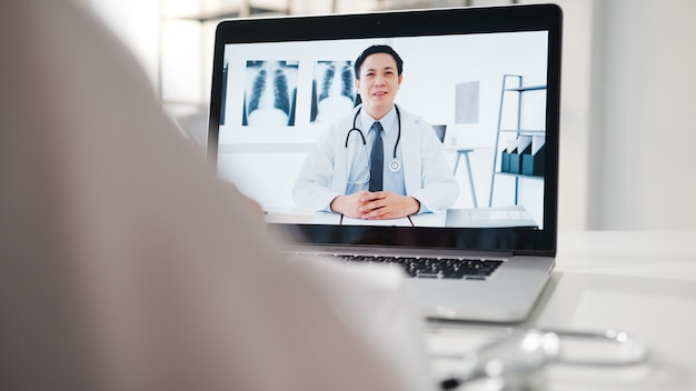 Junge asiatische männliche Ärztin in weißer medizinischer Uniform mit Laptop, die eine Videokonferenz mit einem leitenden Arzt am Schreibtisch in einer Gesundheitsklinik oder einem Krankenhaus spricht.