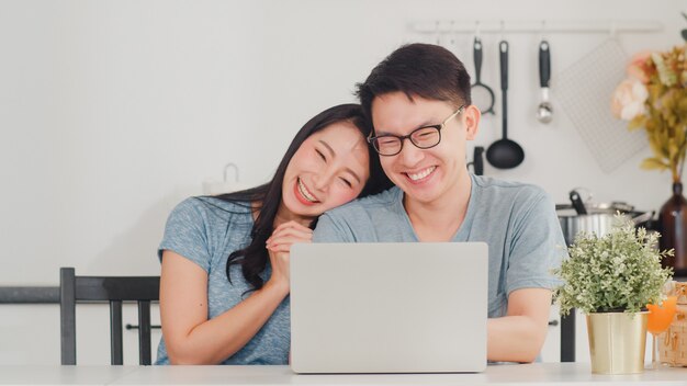 Junge asiatische Paare genießen, online auf Laptop zu Hause zu kaufen. Der junge glückliche Ehemann und die Frau des Lebensstils kaufen elektronischen Geschäftsverkehr, nachdem sie in der modernen Küche am Haus morgens gefrühstückt haben.