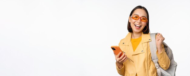 Junge asiatische reisende Mädchentouristin in der Sonnenbrille, die Rucksack und Handy unter Verwendung von appli hält