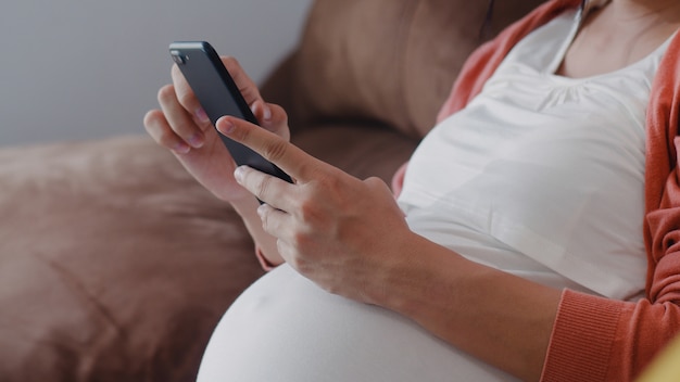 Junge asiatische schwangere Frau, die Handysuchschwangerschaftsinformationen verwendet. Die Mutter, die das glückliche Lächeln positiv und ruhig sich fühlt, während mach s gut ihr Kind, das zu Hause auf Sofa im Wohnzimmer liegt.