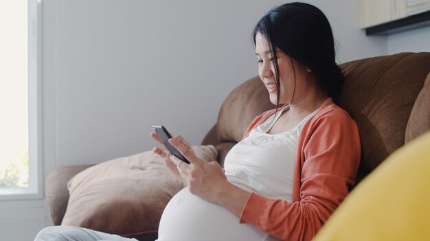 Junge asiatische schwangere Frau, die Handysuchschwangerschaftsinformationen verwendet. Die Mutter, die das glückliche Lächeln positiv und ruhig sich fühlt, während mach s gut ihr Kind, das zu Hause auf Sofa im Wohnzimmer liegt.