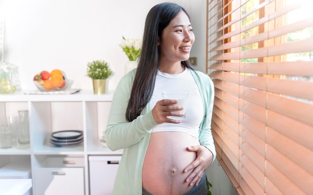 Junge asiatische schwangere Frau, die im Küchenraum steht und ein Glas frische Milch isst, um während der Schwangerschaft bis kurz vor der Geburt eine gute Gesundheit zu erhalten