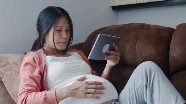 Junge asiatische schwangere Frau, die Tablettensuchschwangerschaftsinformationen verwendet. Die Mutter, die das glückliche Lächeln positiv und ruhig sich fühlt, während mach s gut ihr Kind, das zu Hause auf Sofa im Wohnzimmer liegt.
