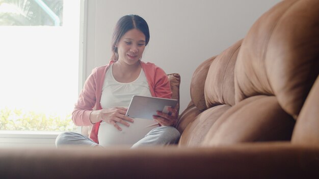 Junge asiatische schwangere Frau, die Tablettensuchschwangerschaftsinformationen verwendet. Die Mutter, die das glückliche Lächeln positiv und ruhig sich fühlt, während mach s gut ihr Kind, das zu Hause auf Sofa im Wohnzimmer liegt.