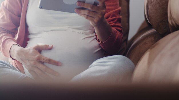 Junge asiatische schwangere Frau, die Tablettensuchschwangerschaftsinformationen verwendet. Die Mutter, die das glückliche Lächeln positiv und ruhig sich fühlt, während mach s gut ihr Kind, das zu Hause auf Sofa im Wohnzimmer liegt.