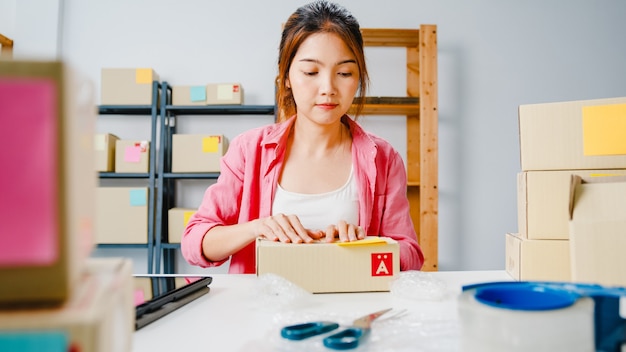Junge asiatische Unternehmer-Geschäftsfrau, die Produkt im Karton verpackt, liefern an Kunden, der zu Hause arbeitet. Kleinunternehmer, Start-up Online-Marktzustellung, Lifestyle freiberufliches Konzept.