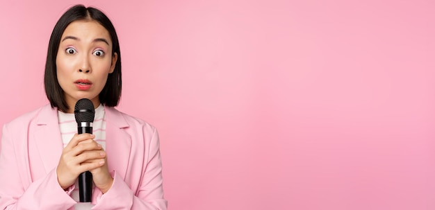 Junge asiatische Verkäuferin im Büro mit Anzug, die ein Mikrofon hält und schockiert in die Kamera schaut, während sie spricht und eine Rede hält, die über rosa Hintergrund steht