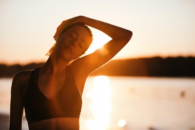 Junge athletische Frau, die ihren Hals streckt, während sie bei Sonnenaufgang am Flussufer trainiert